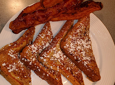 French toast drizzled in powered sugar with a side of bacon served on a white plate